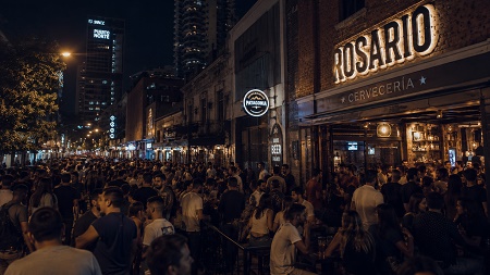 Fotografía panorámica de una concurrida calle peatonal en Rosario de noche, llena de gente en las terrazas de cervecerías con letreros luminosos como "ROSARIO CERVECERÍA".