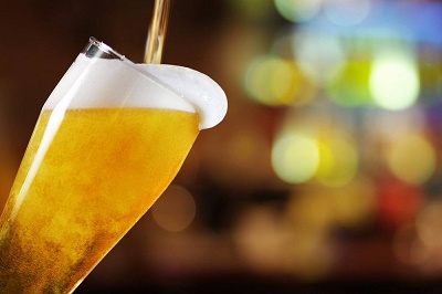 cerveza-tirada-espuma-desborde-primer-plano Primer plano de cerveza tirada con espuma cremosa desbordándose del vaso, líquido dorado ámbar y burbujas en movimiento.